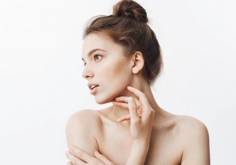 Close up portrait of young female brunette student girl with bun hairstyle and baked shoulders looking aside with calm face expression touching jawline with fingers. Beauty and health